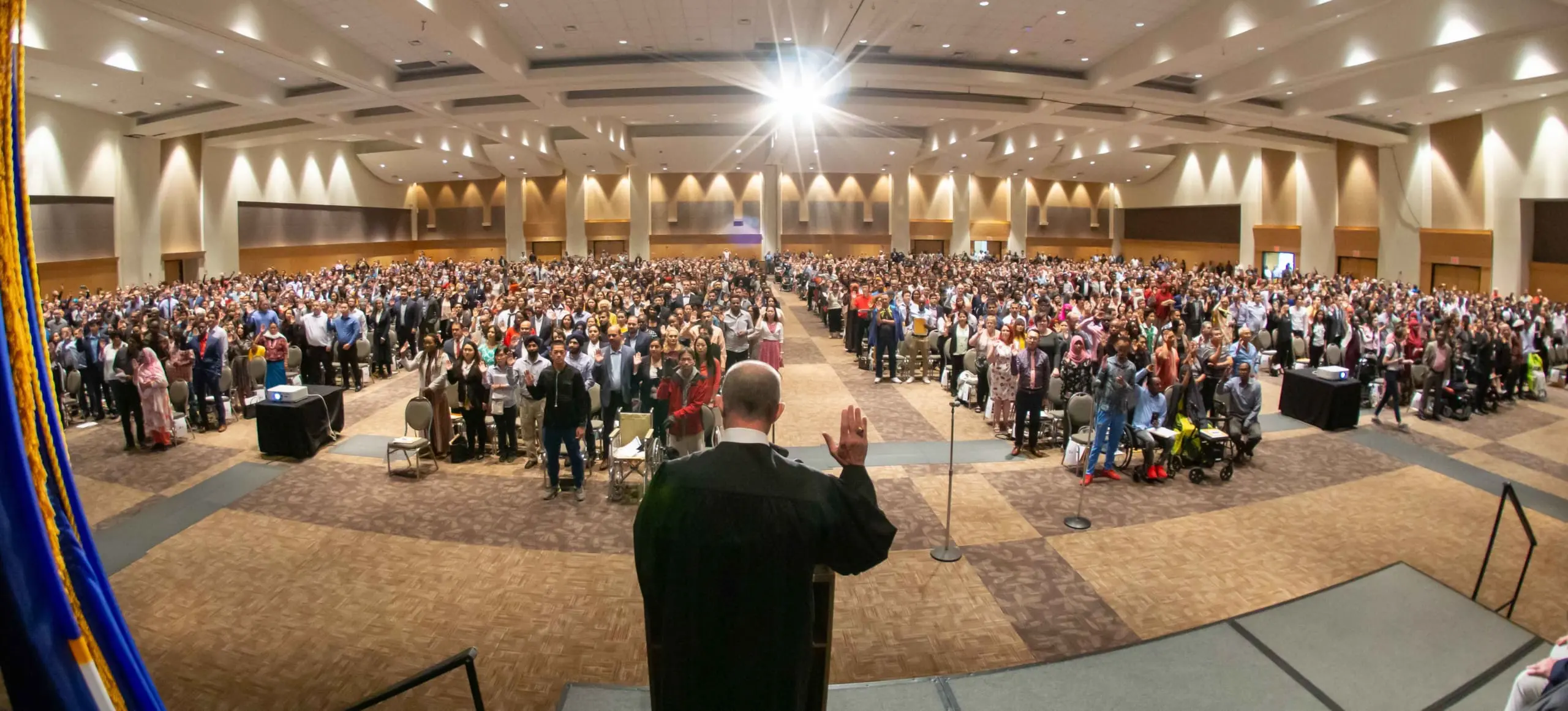 A federal judge with his right hand raised is administering the Oath of Citizenship to a ballroom of new American citizens.